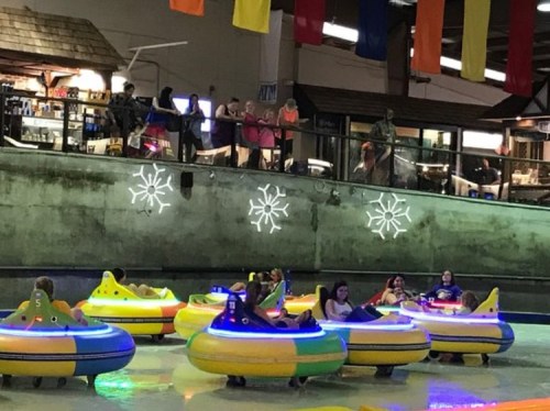 Colorful bumper boats in a water attraction, with people enjoying the ride and spectators watching from above.