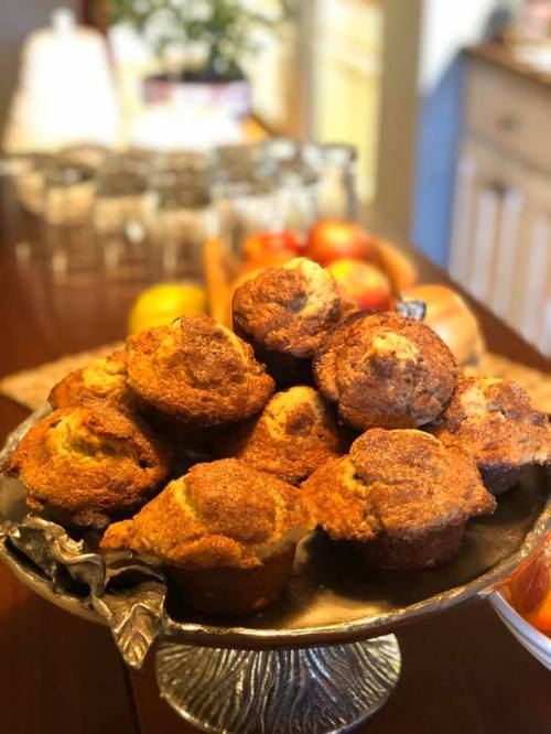 A silver platter piled high with freshly baked muffins, set on a wooden table with fruit and glassware in the background.