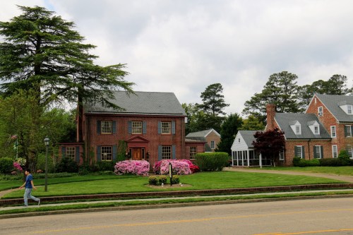 A brick house with colorful flowers in the front yard, surrounded by trees, and a person walking on the sidewalk.