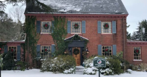 A charming brick house decorated with wreaths, surrounded by snow and greenery, creating a festive winter scene.