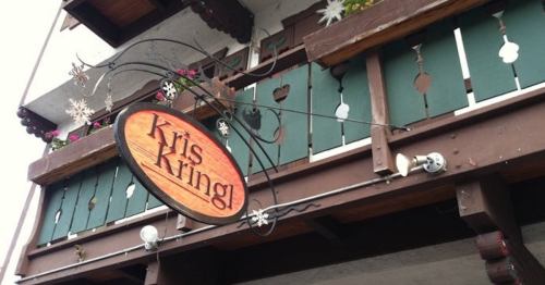 Sign for "Kris Kringl" hanging from a wooden balcony with green shutters and decorative elements.