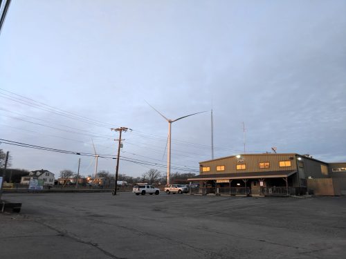 A wind turbine stands beside a building in a quiet area, with power lines and a cloudy sky in the background.