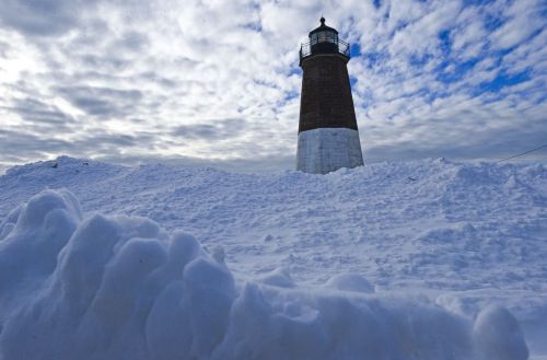 A tall lighthouse stands amidst a snowy landscape under a cloudy sky. Snow drifts surround its base.