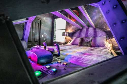 A cozy bedroom with a galaxy-themed wall, featuring a bed and colorful lights, viewed through a unique frame.