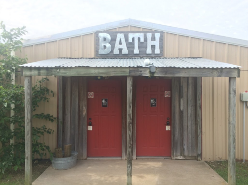 Two red doors labeled "BATH" under a metal roof, surrounded by wooden beams and greenery.