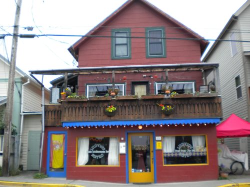 A colorful two-story bistro with a wooden balcony, flower pots, and a bright blue and yellow entrance.