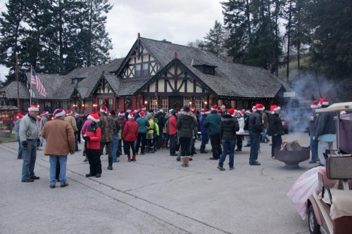 A crowd wearing Santa hats gathers outside a festive building, surrounded by trees and holiday decorations.
