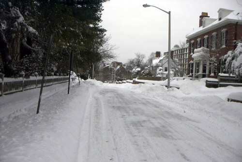 A snow-covered street lined with houses, trees, and a cloudy sky, creating a quiet winter scene.