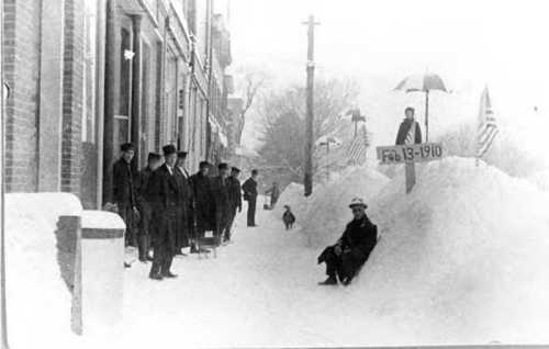 Historic black-and-white photo of men in suits by a snowy street, with a sign dated Feb 13, 1910, and flags in the background.