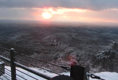 A scenic view of a sunset over a snowy landscape, with mountains in the distance and a cloudy sky.