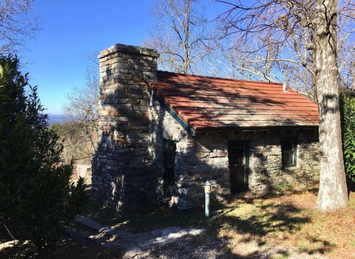 A small stone cabin with a red roof, surrounded by trees and a clear blue sky.