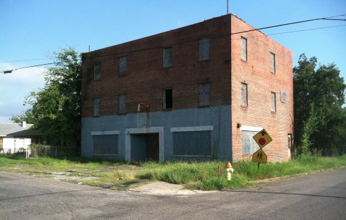 Abandoned brick building with boarded windows, overgrown grass, and a stop sign at the corner of a quiet street.
