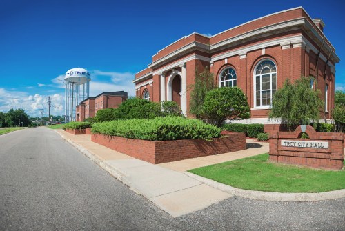 Troy City Hall with a brick facade, landscaped gardens, and a water tower in the background under a clear blue sky.