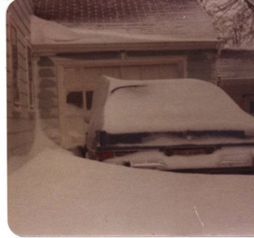 A car buried in snow next to a garage, with heavy snowfall covering the ground and surrounding areas.