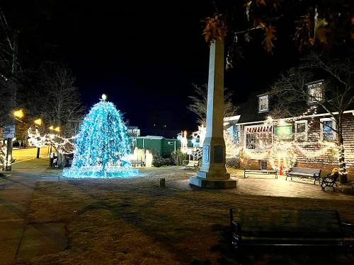 A festive scene at night featuring a lit Christmas tree, decorated buildings, and a monument in a charming town square.