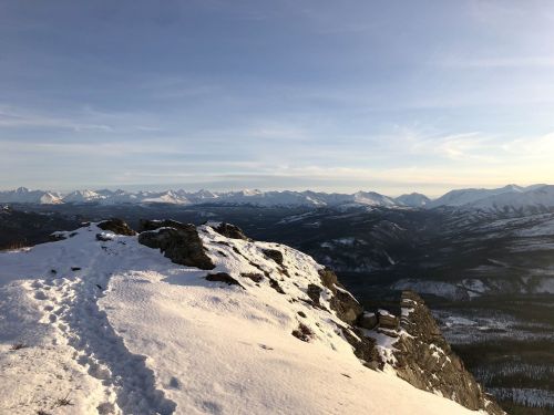 Snow-covered mountain peak with a winding path, overlooking a vast range of mountains under a clear blue sky.