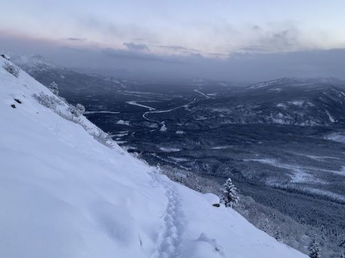 A snowy mountain landscape with a winding river below, surrounded by trees and distant peaks under a cloudy sky.