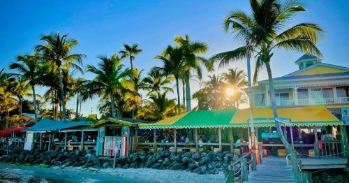 A vibrant beachside restaurant with colorful awnings, surrounded by palm trees and a sunset in the background.