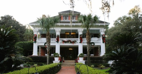 A grand two-story house with a porch, decorated for the holidays, surrounded by lush greenery and palm trees.