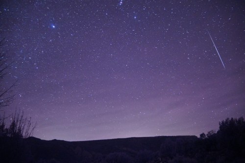A starry night sky filled with stars and a faint meteor streaking across the purple-hued atmosphere.