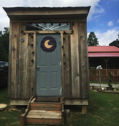 A small wooden shed with a gray door featuring a crescent moon design, set against a blue sky and green grass.