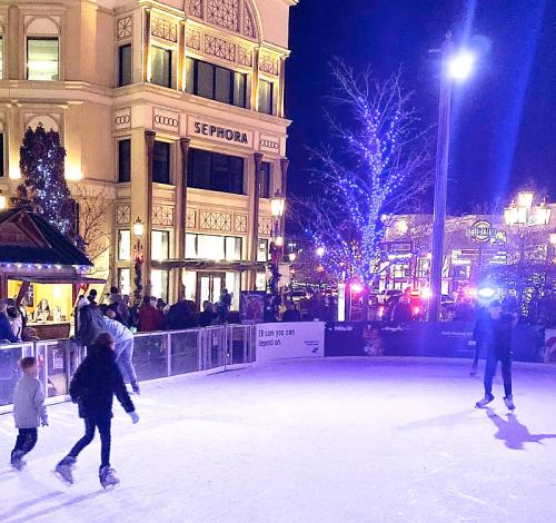 A lively outdoor ice skating rink at night, surrounded by festive lights and shoppers near a Sephora store.