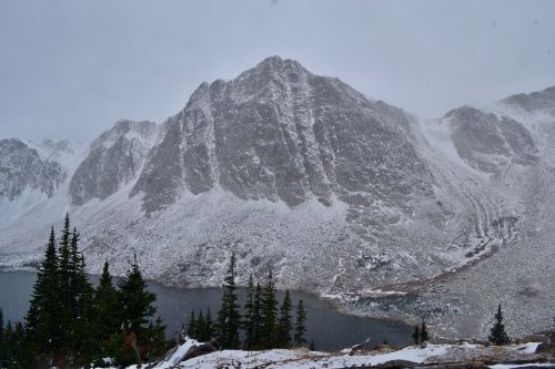 Snow-covered mountain with rugged peaks, surrounded by evergreen trees and a lake, under a cloudy, snowy sky.