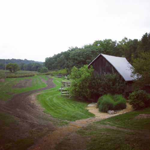 A rustic farm scene with a winding path, green fields, and a weathered barn surrounded by trees under a cloudy sky.