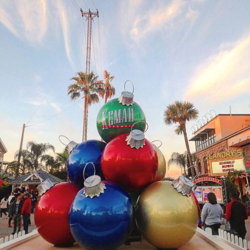 Colorful oversized Christmas ornaments are stacked together, with palm trees and a festive atmosphere in the background.