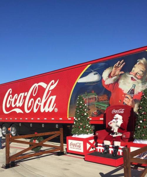 A Coca-Cola truck decorated for the holidays, featuring Santa and Christmas trees against a clear blue sky.