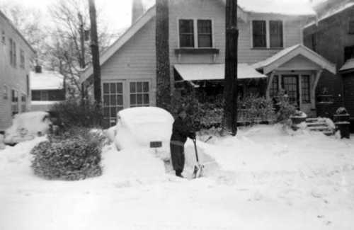 A person shovels snow in front of a house, with heavy snowfall covering the ground and a parked car.