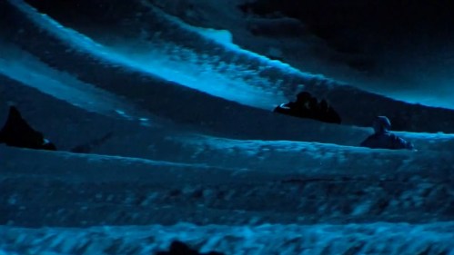 A snowy landscape illuminated by blue light, featuring undulating snowdrifts and silhouettes of people in the distance.