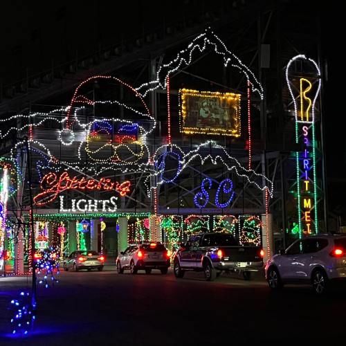 Colorful holiday lights adorn a building and trees at night, with cars parked nearby.