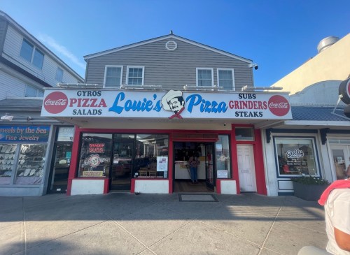 A storefront for Louie's Pizza featuring signs for gyros, salads, subs, grinders, and steaks, with a sunny sky above.