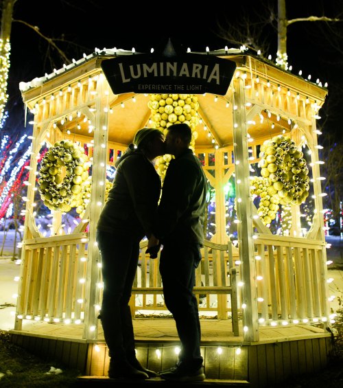 A couple kisses in a beautifully lit gazebo adorned with festive decorations and glowing lights.