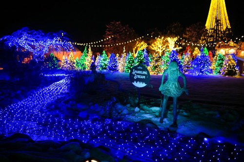 A winter scene with colorful Christmas lights, a sparkling stream sign, and illuminated trees in the background.