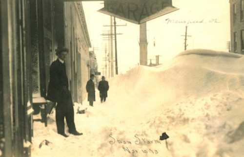 Historic photo of men standing by a snow-covered street, with a garage sign and towering snowdrifts from a severe storm.