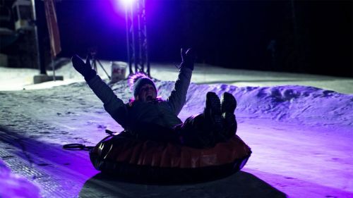 A person joyfully tubing down a snowy slope at night, illuminated by colorful lights.