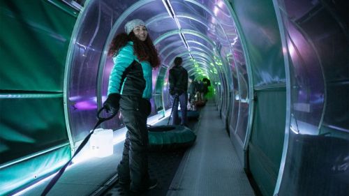 A woman smiles while holding a sled in a brightly lit, colorful tunnel with other people in the background.