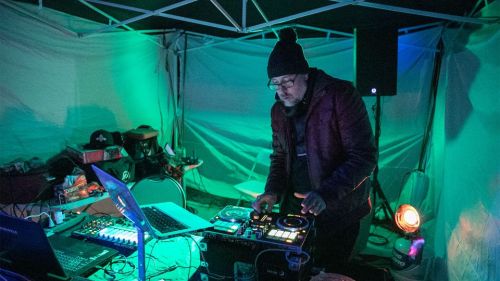 A DJ performs in a dimly lit tent, surrounded by colorful lights and equipment, creating an energetic atmosphere.