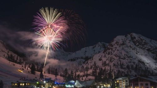 Colorful fireworks illuminate a snowy mountain landscape at night, with buildings in the foreground.