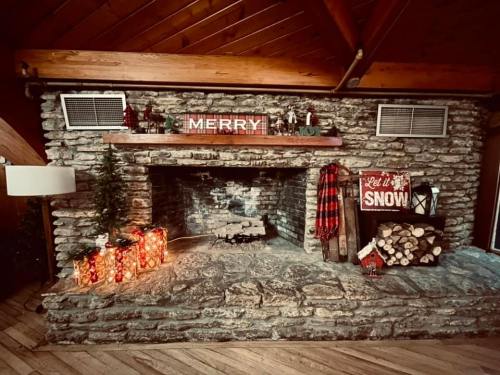 Cozy stone fireplace decorated for the holidays with "MERRY" sign, festive lights, and stacked firewood.
