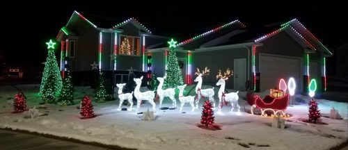 A festive house decorated with colorful Christmas lights, featuring reindeer, a sleigh, and illuminated trees in the snow.