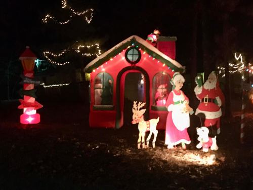 A festive scene with Santa, Mrs. Claus, a reindeer, and a colorful holiday house, all decorated with lights.