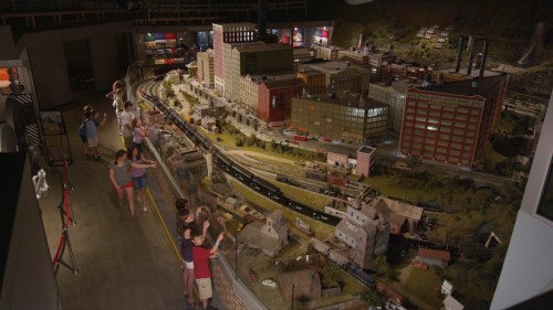 A model train display in a museum, featuring detailed buildings and landscapes, with visitors observing the scene.
