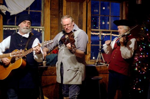 Three musicians perform indoors, playing guitar, violin, and fiddle, with a decorated Christmas tree in the background.