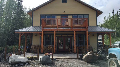 A two-story yellow house with a wooden porch, surrounded by trees and rocks, featuring a bike and flower beds in front.