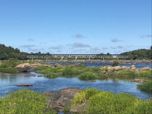 A serene river scene with rocks and greenery, featuring a bridge in the background under a clear blue sky.