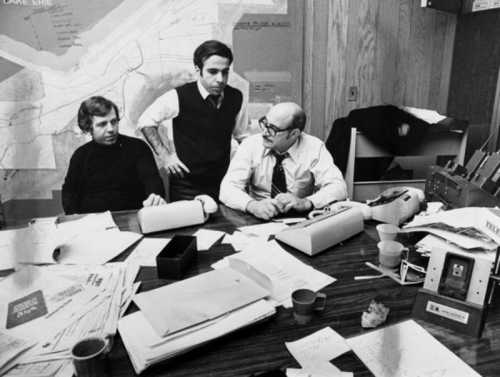 Three men discuss plans at a cluttered table covered with papers and maps in a dimly lit office.