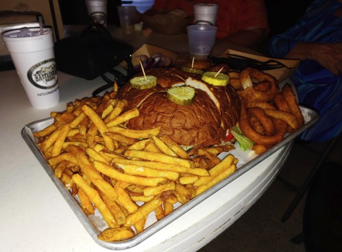 A large burger topped with pickles, surrounded by golden fries and onion rings on a tray.